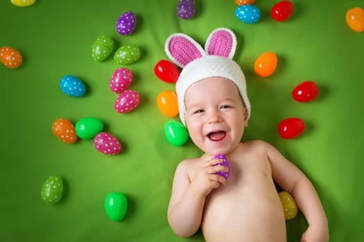 Baby boy in bunny hat lying on green blanket with multicolored easter eggs