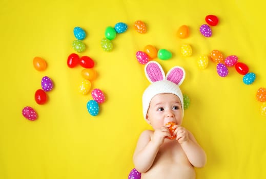 Baby boy in bunny hat lying on yellow blanket with multicolored easter eggs