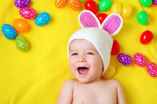 Baby boy in bunny hat lying on yellow blanket with multicolored easter eggs