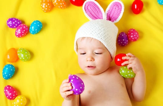 Baby boy in bunny hat lying on yellow blanket with multicolored easter eggs