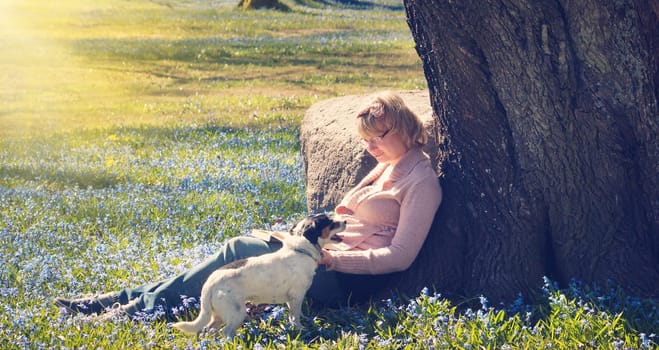 Young woman reading a book with dog under a tree in early spring. Relaxed women in beautiful morning in park with springflowers