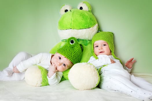 two cute six month old babies lying in frog hats with a soft toy