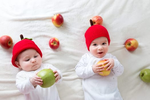 two cute six month old babies lying in hats on soft blanket with apples