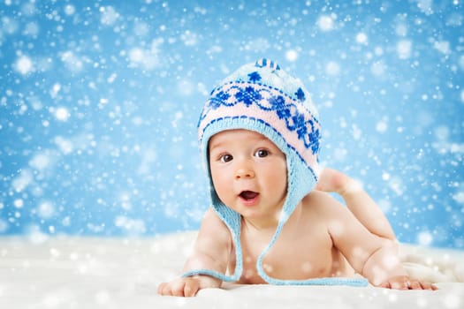 Eight month old baby in knitted winter hat lying on soft blanket with blue background and falling snow
