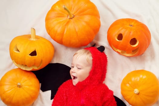 Baby in devil costume with pumpkins. Child in halloween outfit lying on soft white blanket