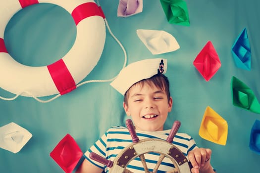 seven years old boy playing in sailor hat. Child with a steering wheel and paper ships on blue background