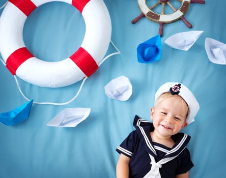 two years old boy playing in sailor hat. Child with a steering wheel and paper ships on blue background