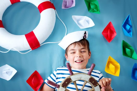 seven years old boy playing in sailor hat. Child with a steering wheel and paper ships on blue background