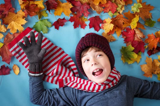 seven years old boy dreaming in autumn. Child in hat and scarf and maple leaves on blue background