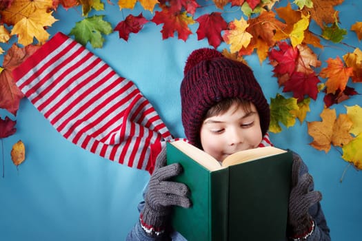 seven years old boy dreaming in autumn. Child in hat and scarf and maple leaves on blue background reading a book