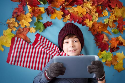 seven years old boy dreaming in autumn. Child in hat and scarf and maple leaves on blue background holding a tablet