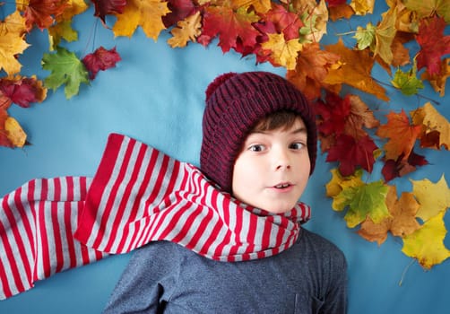 seven years old boy dreaming in autumn. Child in hat and scarf and maple leaves on blue background