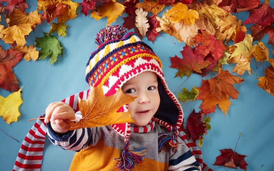 two years old boy dreaming in autumn. Child in hat and scarf and maple leaves on blue background