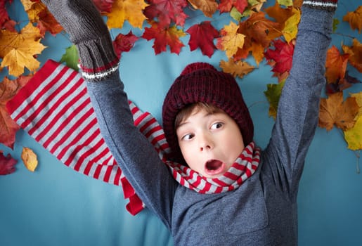 seven years old boy dreaming in autumn. Child in hat and scarf and maple leaves on blue background