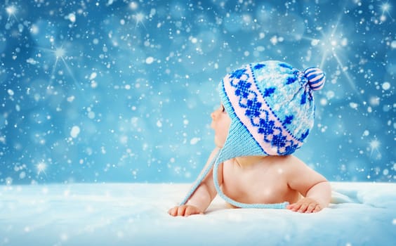 Eight month old baby in knitted winter hat lying on soft blanket with blue background and falling snow