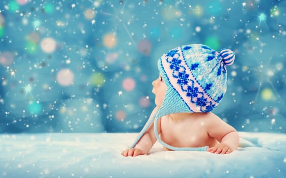 Eight month old baby in knitted winter hat lying on soft blanket with blue background and falling snow