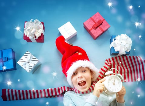 Happy child on blue blanket in red hat with alarm clock. Cute boy in christmas cap with many presents