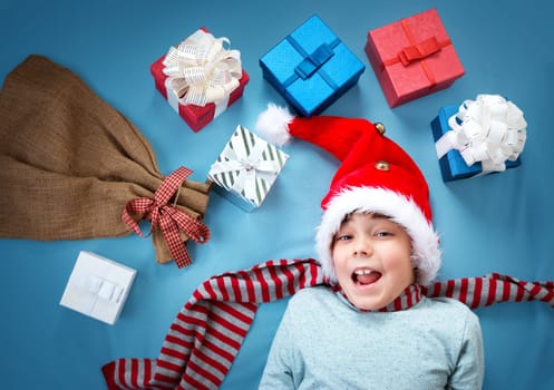 Happy child on blue blanket in red hat holding gift box. Cute boy in christmas cap with many presents