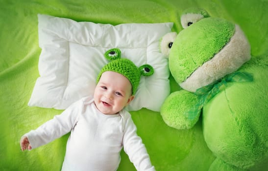 little boy in knitted animal hat lying on soft green blanket with frog softtoy. Cute 5 month old baby on the pillow in bed