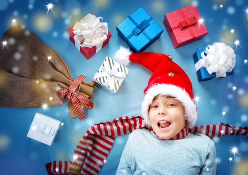 Happy child on blue blanket in red hat holding gift box. Cute boy in christmas cap with many presents