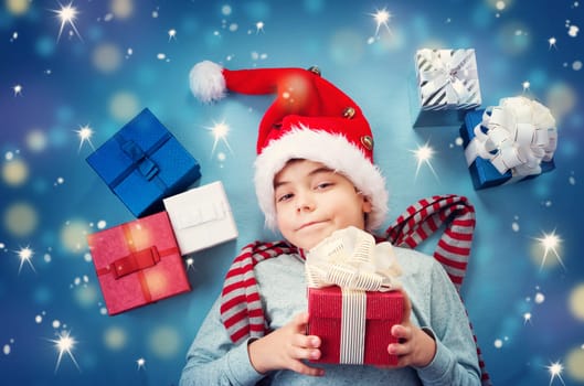 Happy child on blue blanket in red hat holding gift box. Cute boy in christmas cap with many presents