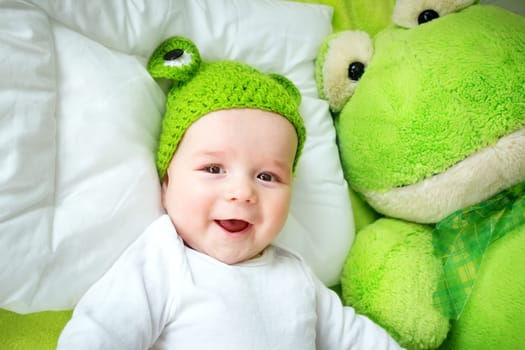 little boy in knitted animal hat lying on soft green blanket with frog softtoy. Cute 5 month old baby on the pillow in bed