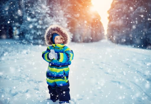Baby boy walking at snowfall. Child in winter clothes playing with snow