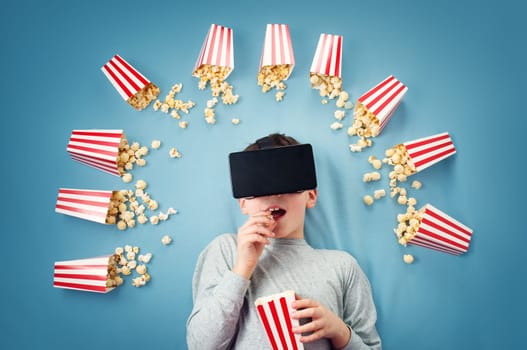 Child lying on blue background in Virtual reality glasses and watching film. Boy eating popcorn