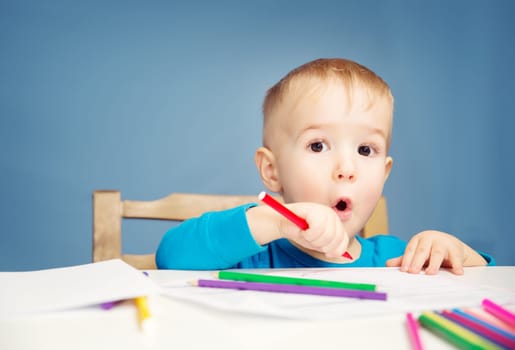 Little child drawing on the paper. Boy sitting on the chair at table with pencils