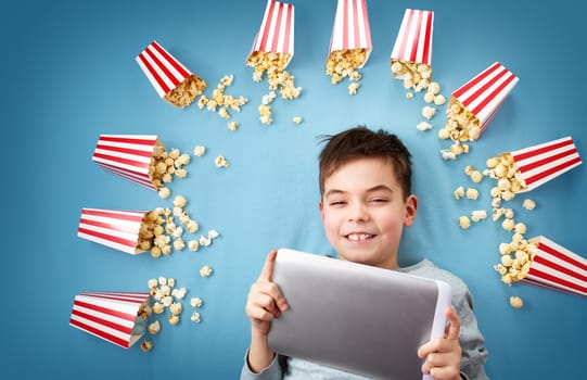 Child lying on blue background with tablet and watching movie. Boy with popcorn