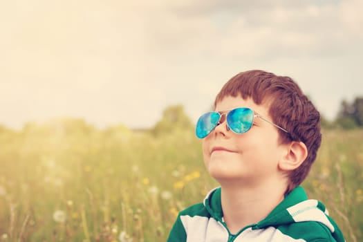 Happy child in sunglasses on the field with dandelions. Boy looking to the sky in summer outdoors