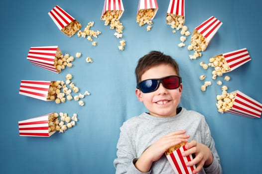 Child lying on blue background in 3d glasses and watching tv. Boy eating popcorn