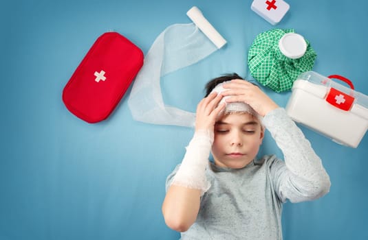 Sad child in bandages lying in bed. Wounded boy on blue background with first aid accessories.