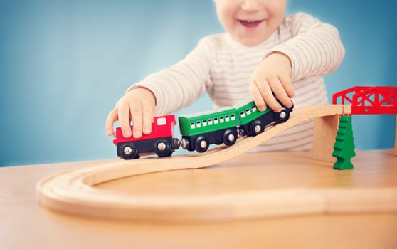 Portrait of a two years old child sitting at the table and playing with a railroad. Pretty little boy on blue background