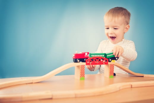 Portrait of a two years old child sitting at the table and playing with a railroad. Pretty little boy on blue background