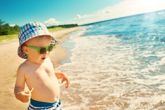 one year old boy walking at the beach in summer hat and sunglasses. Child on family vacations at sea