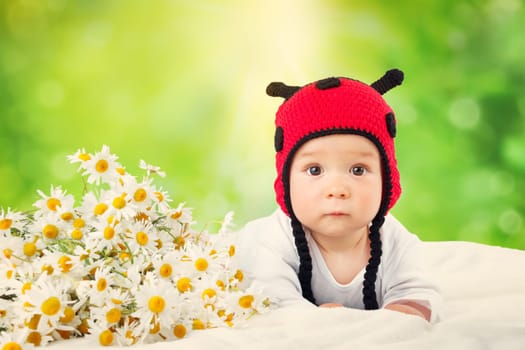 Cute baby lying in the bed on white blanket in ladybug hat. Little boy with chamomille flowers