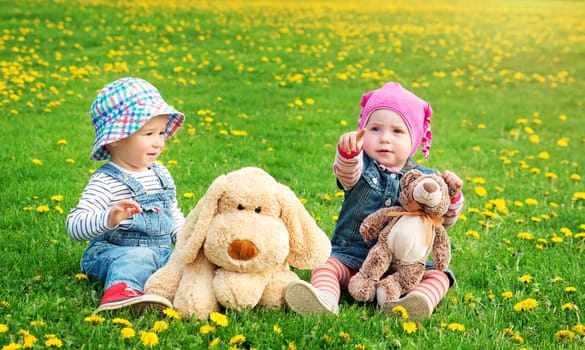 Little boy and girl in hats sitting on the field with soft toys in summer. Two cute one year old babies on the grass with dandelions