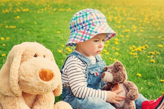 sad boy sitting with softtoys outoors. Thoughtful child with teddy and plush dog on the field with dandelions in spring