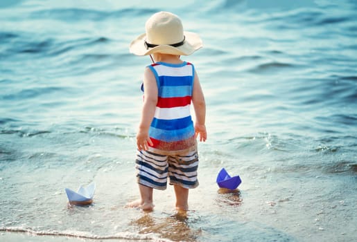 one year old boy playing at the beach in straw hat. Child with a paper ship at sea