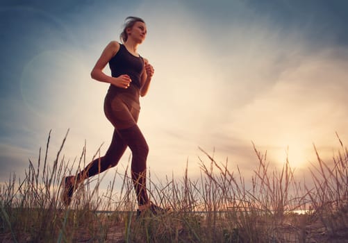 Young woman running on the field near seaside at sunset. Active person outdoors at the dusk in summer