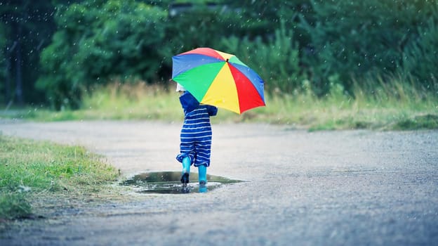 Child walking in wellies in puddle on rainy weather. Boy holding colourful umbrella under rain in summer