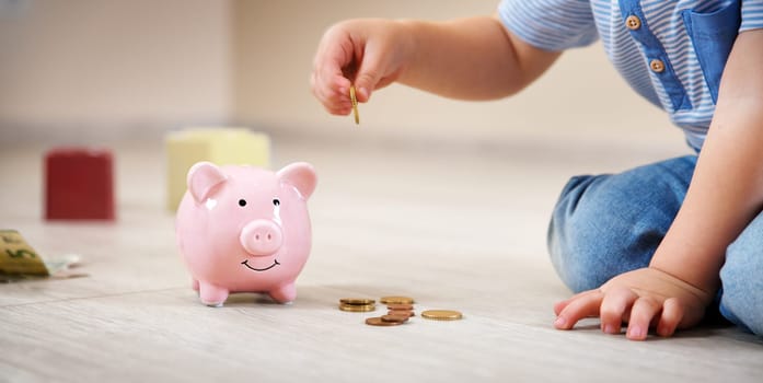 two years old child sitting on the floor and putting a euro coin into a piggybank.