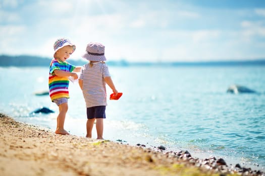 Babygirl and babyboy standing on the beach in summer hats and holding paper ships