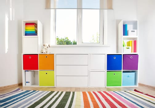 Colourful children rooom with white walls and furniture. Rainbow carpet at home interior with a window