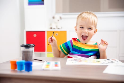 Little child drawing on the paper. Boy sitting on the chair at table with paints