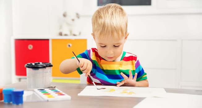 Little child drawing on the paper. Boy sitting on the chair at table with paints