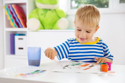 Little child drawing on the paper. Boy sitting on the chair at table with paints