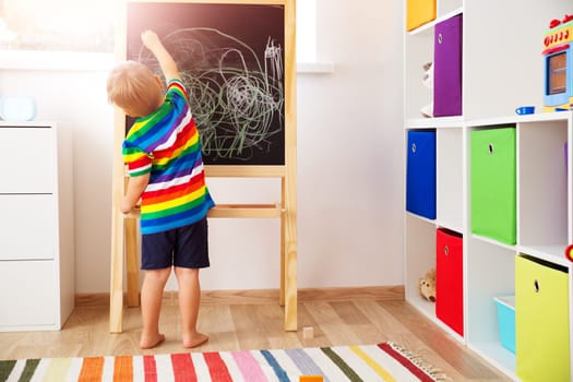 Little child drawing on the blackboard. Boy standing in the room with chalkboard