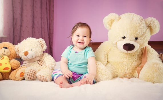 Cute smiling little baby girl sitting on the bed with soft toys. Seven month old infant child on white soft blanket